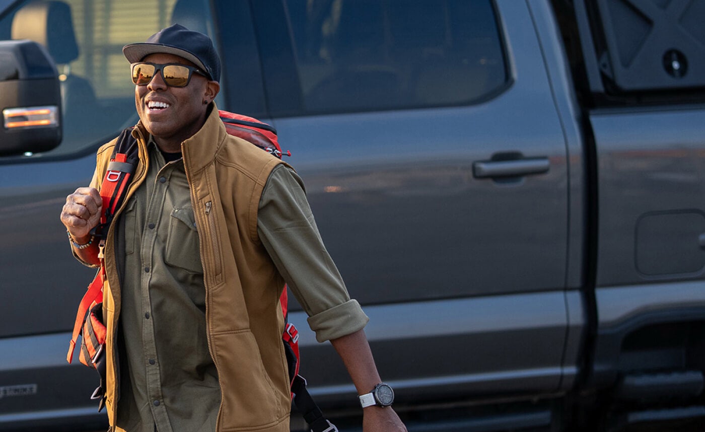 A smiling man with a backpack slung over one shoulder walking away from his grey Ford F-150.