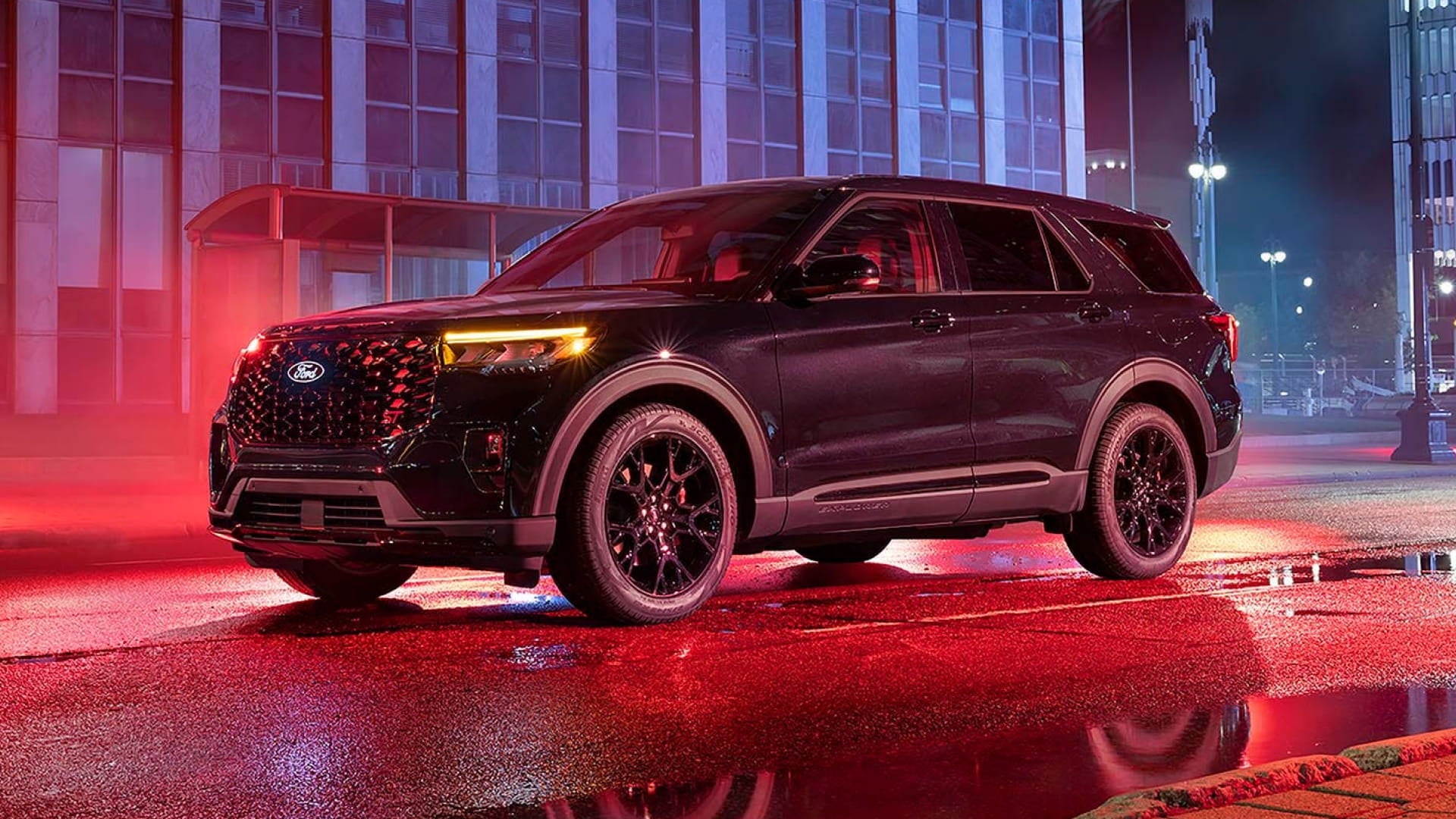 Front three-quarter view of a black Ford Explorer with illuminated headlights on a wet city street under red and blue lighting at night.