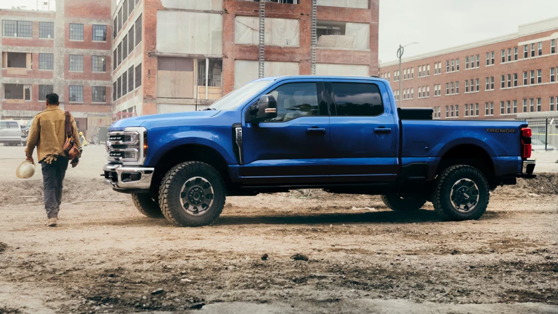 Side view of a blue Ford Super Duty F-250 truck parked at a construction site as a worker walks nearby carrying equipment.