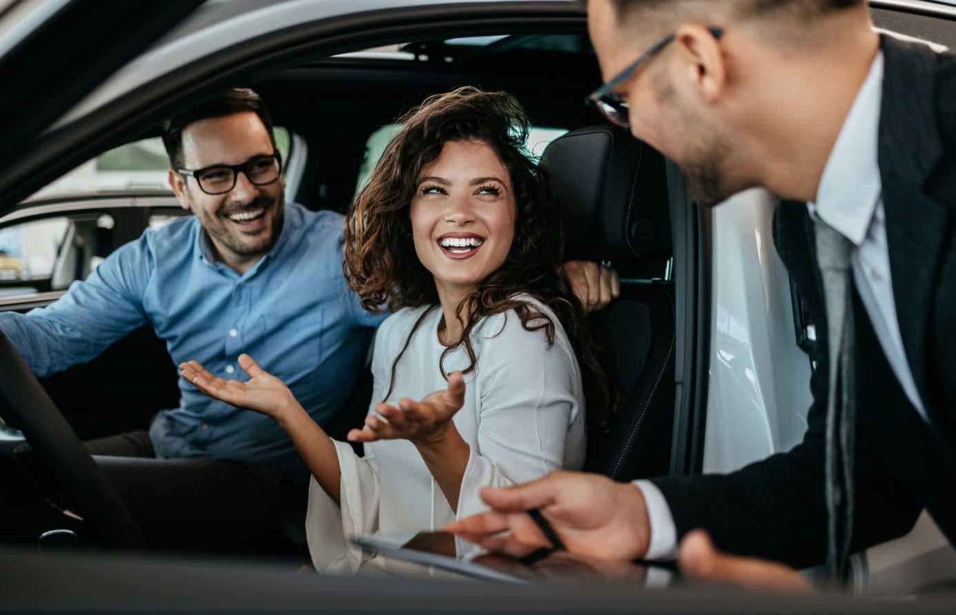 Smiling couple sitting inside a vehicle while speaking with a salesperson.